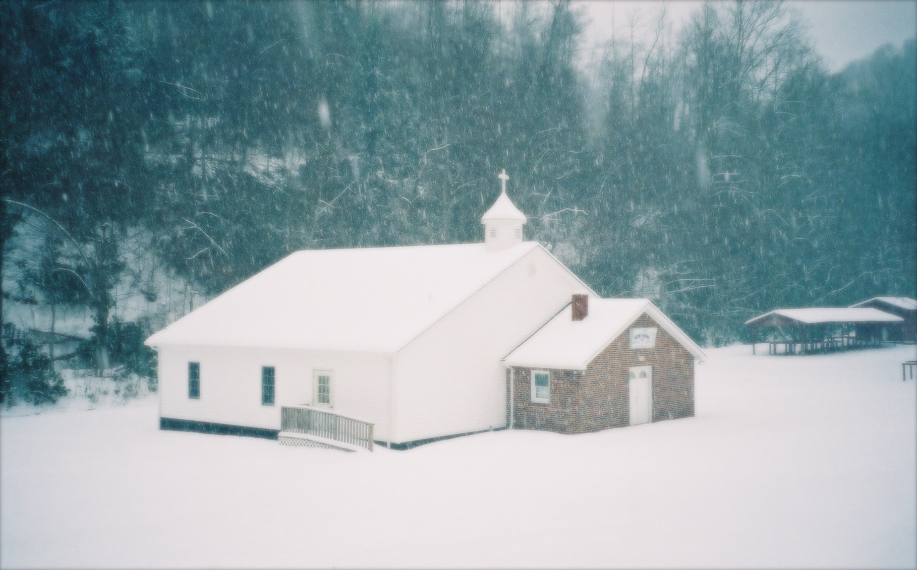 White country church and stone fellowship hall surrounded by unbroken snow during active snowfall, NC High Country